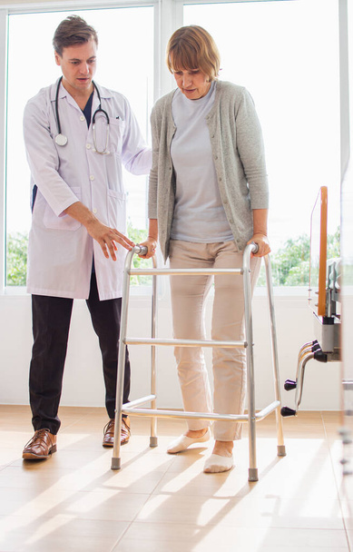 Blanke mannelijke arts helpt vrouwelijke patiënt proberen te lopen met walker. De dokter onderzoekt de patiënt in het ziekenhuis. - Foto, afbeelding