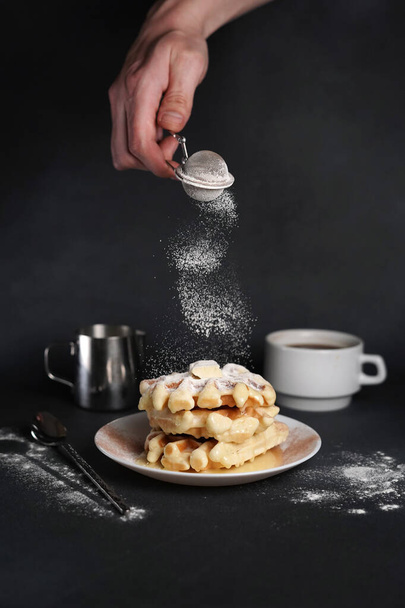 Kaukázusi Man 's Hand megszórja cukor por finom gofri, karamell szósz, kávé csésze, tej, desszert kanál, szűrő fekete beton háttér - Fotó, kép