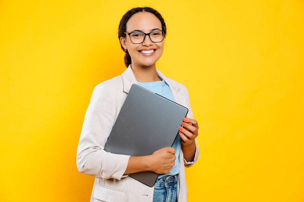 Photo of a successful confident brazilian or hispanic business lady in stylish elegant clothes, creative manager, holding laptop, stand on isolated yellow background, looks at camera, smiles friendly - Foto, afbeelding