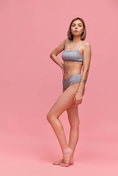 Portrait of young thin sportive woman in grey cotton inner wear posing over pink background. Concept of natural beauty, body and skin care, health, anti-cellulite program, ad. - Zdjęcie, obraz