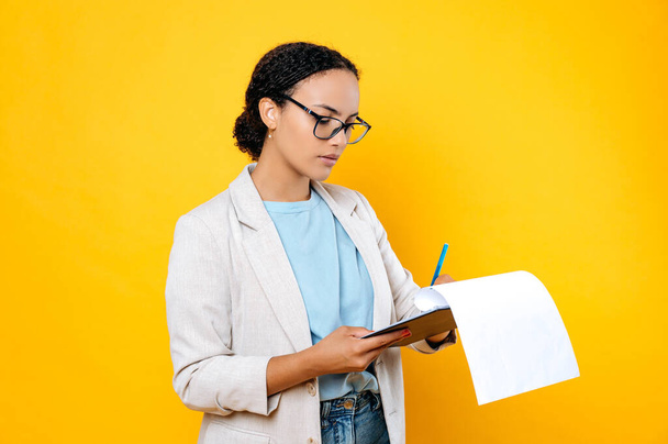 Focused successful brunette mixed race woman, wearing stylish casual clothes, hr manager, real estate agent, holds and sign documents, concentrated looks at them, stand on isolated yellow background - Photo, Image
