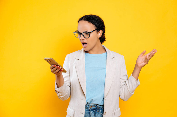 Irritated angry brazilian or latino woman using her smartphone, looking at the screen with displeasure, outraged by the message or news, gesturing with her hand, stand on isolated yellow background - Photo, image