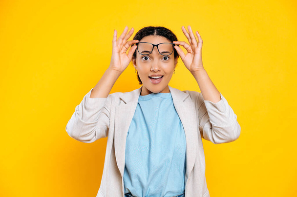 Surprised stunned joyful mixed race woman in stylish elegant clothes, business lady, looking at the camera in amazement, taking off glasses from her eyes, stand on isolated yellow background, smiling - Foto, afbeelding
