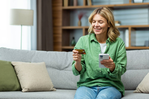 Smiling senior beautiful woman sitting on sofa at home. Holding a phone, using a credit card. Smiling makes online purchases, opened an account, checks. - Photo, Image