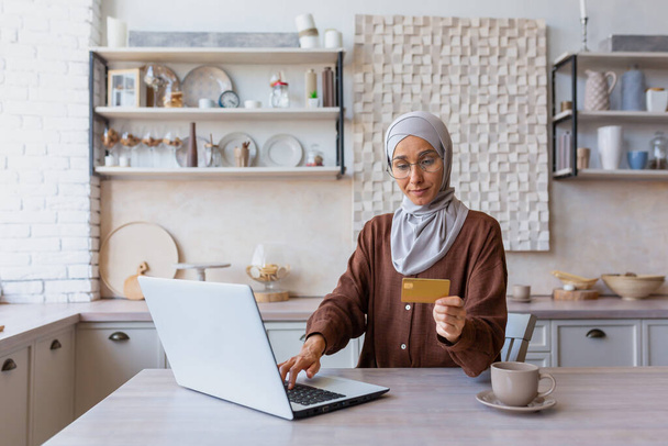 Giovane donna musulmana in hijab che lavora in remoto online. Seduto in cucina digitando su un computer portatile e utilizzando una carta di credito. Acquisti online, pagamenti, transazioni, investimenti, conti. - Foto, immagini