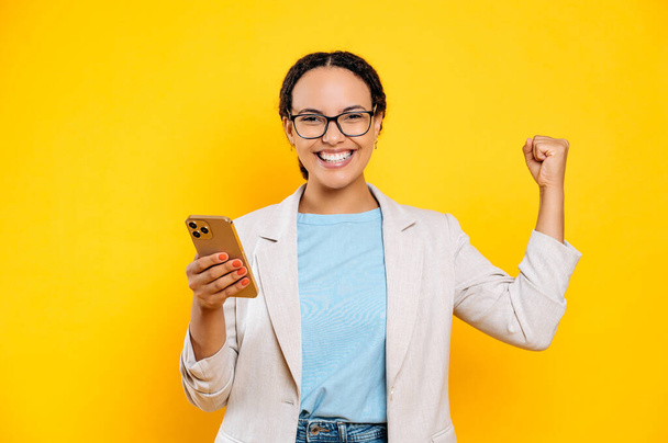 Радісна щаслива змішана жінка в окулярах, тримає свій смартфон в руці, радіє успіху або перемозі, дивиться на камеру, робить жест YES, стоїть на ізольованому жовтому тлі
 - Фото, зображення