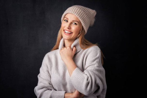 Beautiful smiling woman standing at isolated dark background. Blond haired woman wearing knit hat and turtleneck sweater. Copy space.  - Фото, изображение