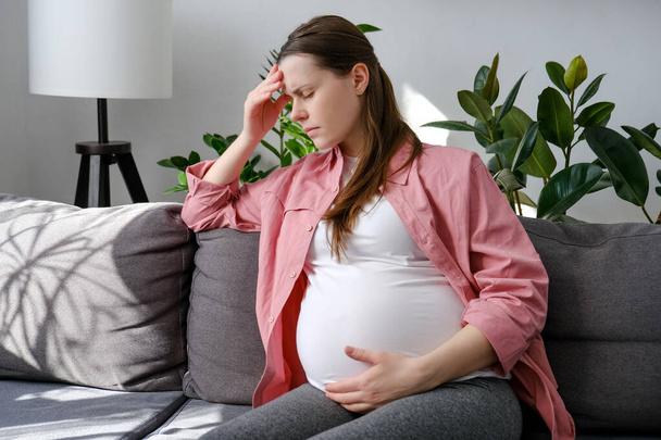 Worried young pregnant female feeling stress and anxiety while sitting alone on couch in living room at home. Expectant mother thinking about upcoming birth of child. Depression and motherhood concept - Photo, image
