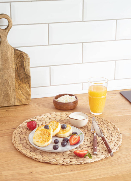 Чизкейки или творог со свежими ягодами, сметаной и апельсиновым соком на кухонном столе. Концепция здорового и вкусный завтрак. - Фото, изображение