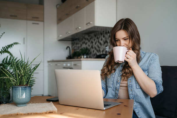 Mulher bonita desfrutando de uma xícara de café enquanto relaxa com seu laptop na cozinha. A trabalhar em casa em quarentena. Distanciamento social Auto-isolamento. Foto de alta qualidade - Foto, Imagem