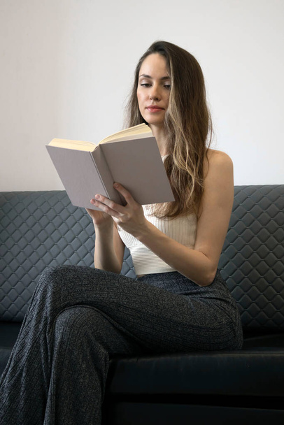 Kobieta w bieli i szarości siedzi na kanapie i czyta książkę. Kobiety w literaturze. - Zdjęcie, obraz