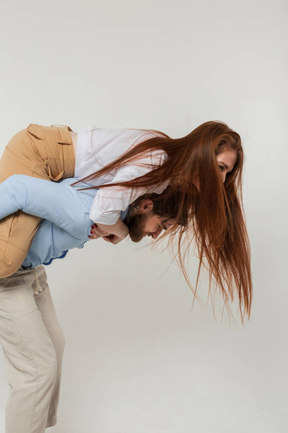 Beautiful young couple man and redheaded girl fooling around and having fun on a white background in the studio. Woman on a guy's back - Fotó, kép