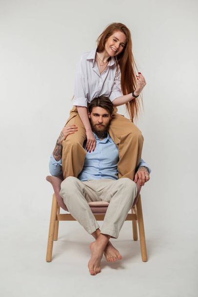 Happy funny fashion couple, beautiful cheerful woman sit on shoulders of hipster brutal guy in fashion clothes on white background in studio - Photo, image
