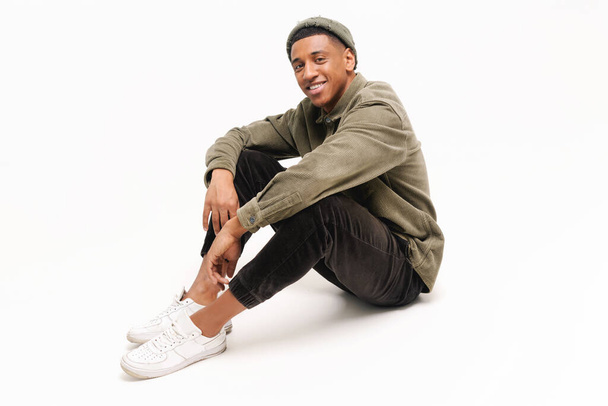 Full length photo of carefree smiling african-american guy in hat sitting on the floor in relaxed pose isolated on white background - Foto, immagini