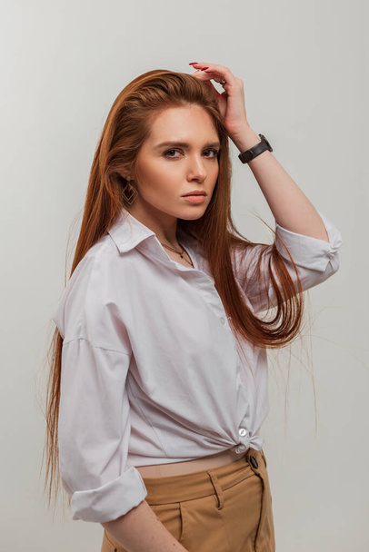 Όμορφη κοκκινομάλλα κοπέλα με ένα χαριτωμένο πρόσωπο σε ένα μοντέρνο λευκό πουκάμισο στέκεται στο στούντιο σε λευκό φόντο - Φωτογραφία, εικόνα