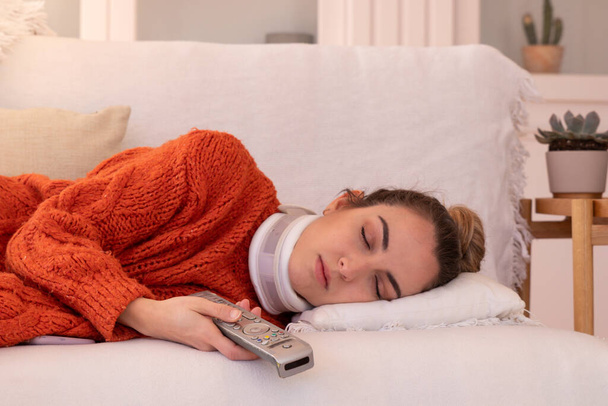Mujer joven en suéter naranja con cuello ortopédico que sostiene el control remoto de la TV mientras duerme en un cómodo sofá en la sala de estar en casa - Foto, imagen