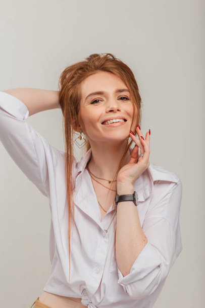 Χαριτωμένο γυναικείο πορτρέτο μιας όμορφης νεαρής κοκκινομάλλας με κόκκινα μαλλιά με ένα όμορφο λευκό χαμόγελο σε λευκό πουκάμισο σε λευκό φόντο στο στούντιο - Φωτογραφία, εικόνα