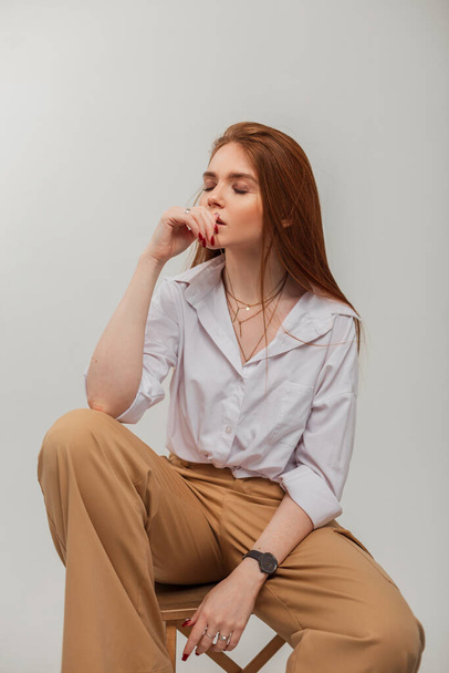 Smyslná jemná krásná zrzavá dívka s dlouhými zrzavými vlasy v elegantním oblečení s bílou košilí a kalhotami sedí na dřevěné židli - Fotografie, Obrázek