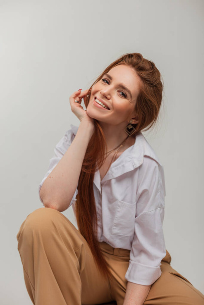 Natürliche weibliche Porträt einer schönen fröhlichen netten Frau mit einem schönen Lächeln in Modekleidung mit Hose und Hemd sitzt auf weißem Hintergrund im Studio - Foto, Bild