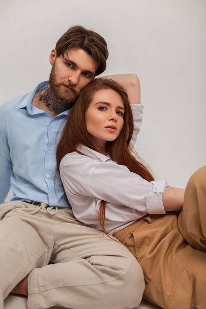 Kaunis muodikas nuori pari trendikkäissä tyylikkäissä vaatteissa istuu ja poseeraa studiossa valkoisella taustalla - Valokuva, kuva