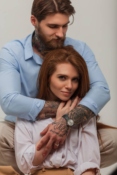Fashion portrait of a beautiful stylish couple in the studio. Handsome brutal man with a beard and a tattoo hugs a beautiful happy fresh red-haired girl - Fotoğraf, Görsel