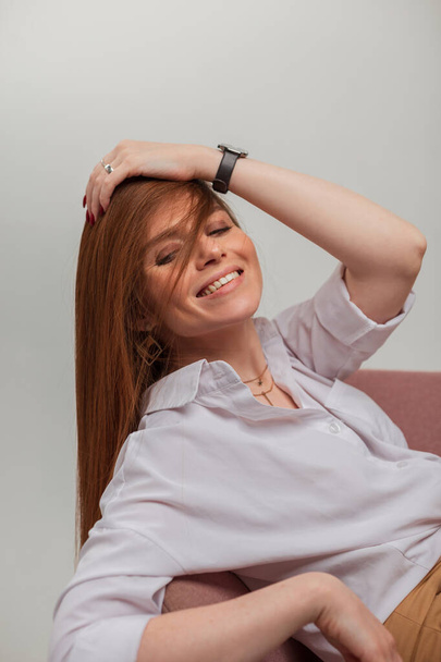 Moda beyaz tişörtlü güzel, güzel, kızıl saçlı kız bir koltuğa oturur ve stüdyoda eğlenir. - Fotoğraf, Görsel
