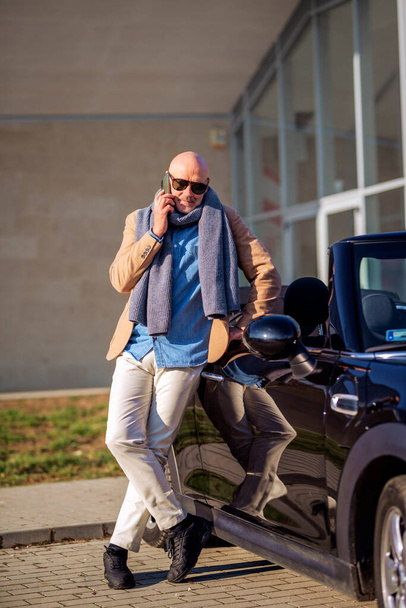 Full length shot of middle aged man wearing casual clothes and using mobile phone while standing outdoor beside his car. - Foto, imagen