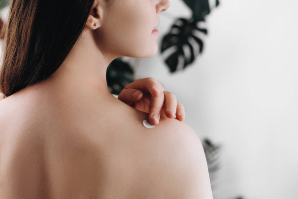 Close up back view of young female 20s old years applying cream on shoulder standing in cozy light bathroom at home. Beautiful lady apply body lotion. Health and beauty concept. Daily skincare routine - 写真・画像