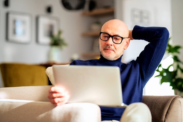 Mid aged man looking thoughtfully while sitting at home and using laptop for work. - 写真・画像