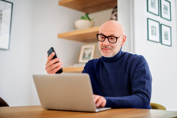 Чоловік середнього віку використовує ноутбук і смартфон під час роботи з дому. Впевнений чоловік сидить за столом вдома. Домашній офіс
. - Фото, зображення