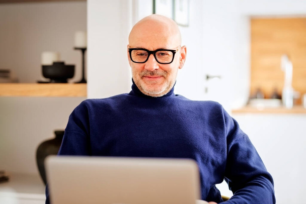 Middle aged man using laptop and having video call while working from home. Confident male sitting at table at the kitchen. Home office. - Photo, Image