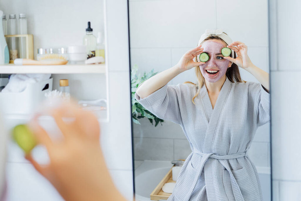 A young smiling woman with pink clay facial mask holds cucumber slices making a refreshing eye mask in bathroom. Natural cosmetic procedures for skin care at home. Beauty self-care. Selective focus. - Foto, Bild