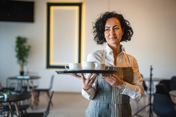 Eine reife Frau kaukasischen Kellnerin im Café oder Restaurant tragen Tablett mit Kaffee weibliche Unternehmerin bei der Arbeit echte Menschen kopieren Raum kleines Geschäftskonzept - Foto, Bild