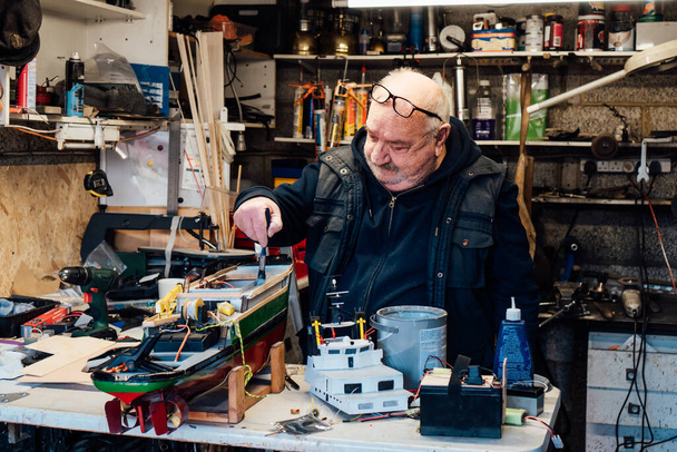 Senior man, pensioner making ship model building handcraft on table with different materials in his garage. Retired elderly man painting boat part. Active Retirement lifestyle, hobby. Selective focus - Photo, Image