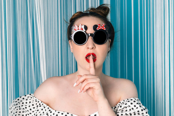 Женщина с ярко-красными раскрашенными губами, одетая в черно-белое платье в горошек и черные круглые солнцезащитные очки с плюшевой игрушкой на белом и синем занавесках. Стиль 50-х. - Фото, изображение