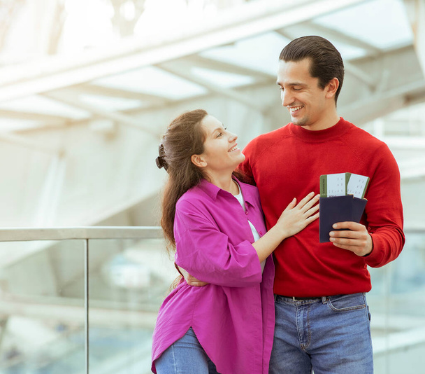 Cheap Tickets. Happy Travelers Couple Holding Passport And Boarding Passes Hugging Standing In Modern Airport Terminal Indoors. Millennial Spouses Going On Vacation Together - Foto, Imagem