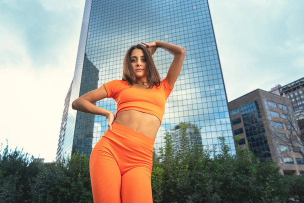 Photo shoot of stylish caucasian hip hop dancer posing having fun in orange leggings and top gymnastic jumpsuit, enjoy the urban town as background. City buildings shooting of moving fitness model - Foto, Imagen
