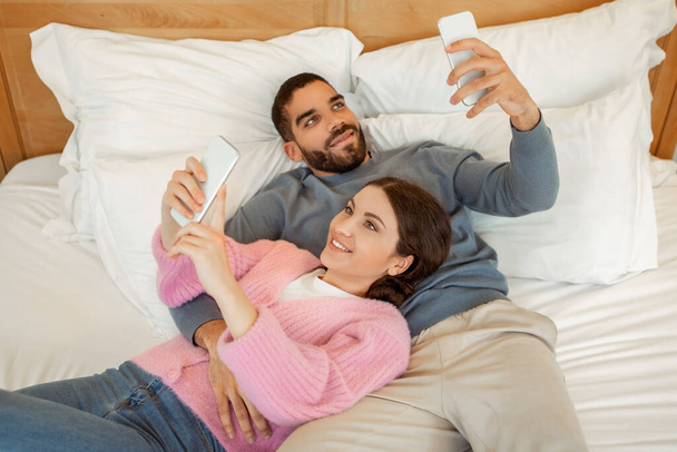 Para za pomocą smartfonów SMS-y i przeglądanie Internetu leżącego w łóżku w nowoczesnej sypialni w domu. Milenijni małżonkowie korzystający z nowej aplikacji telefonicznej. Koncepcja technologii i gadżetów. Powyżej widoku - Zdjęcie, obraz