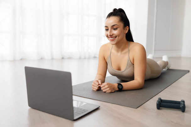 Online fitness class concept. Attractive brunette cheerful sporty woman exercising at home, lying on yoga mat on her belly, using laptop, enjoying newest online pilates course, copy space - Photo, Image