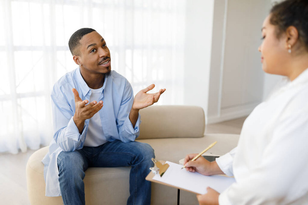 Handsome millennial black guy in casual sitting on couch at therapist office, sharing feelings and gesturing, young anxious man attend therapy session with mixed race woman psychologist, free space - Photo, image
