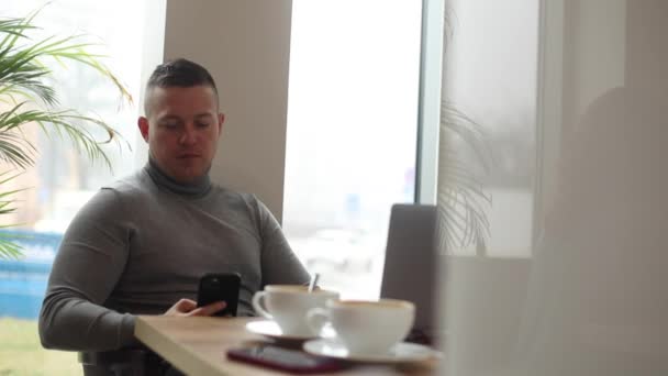 Красивий чоловік з невдоволенням надсилає смс на смартфон і п'є каву в кафе. Бізнесмен працює на ноутбуці, сидячи за столом у кав'ярні. Робочий спосіб життя, ділова пошта, кава-брейк
. - Кадри, відео