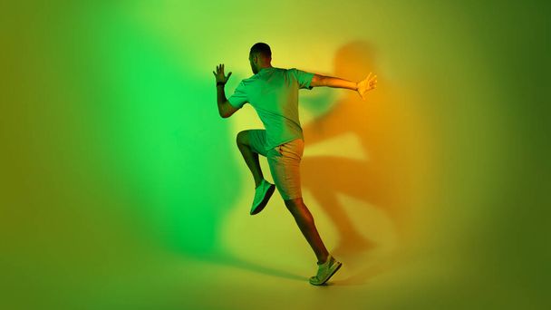 Afroamerykanin Mężczyzna Running Training Over Green Neon Studio Tło. Pełna długość ujęcia nierozpoznawalnego sportowca noszącego odzież sportową. Panorama, widok z boku - Zdjęcie, obraz