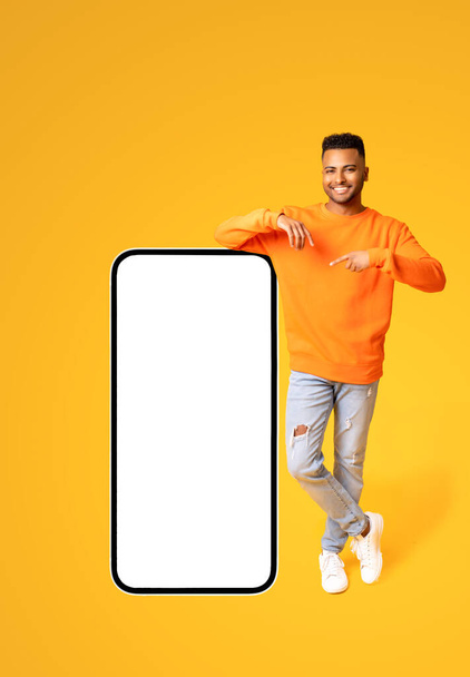 Playful handsome indian man pointing finger at huge smartphone with empty blank screen isolated on yellow, happy arab guy presenting mobile app, showing best deal, copy space, template - Fotó, kép