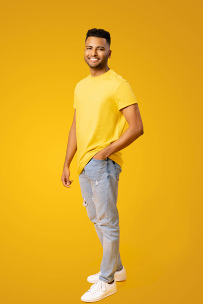 Portrait of happy satisfied Indian young man in t-shirt standing and looking at the camera with light smile isolated on yellow. Indoor studio shot of handsome guy in full length - Fotografie, Obrázek