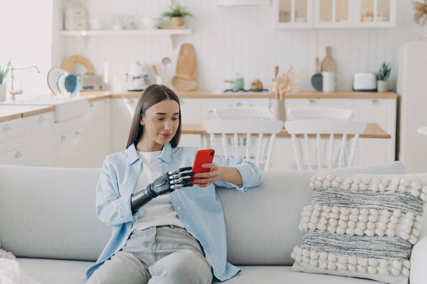 Attractive handicapped woman is texting on smartphone. Disabled european woman browsing internet. Happy girl is holding the phone with bionic artificial arm. Equality and life quality concept. - Photo, Image