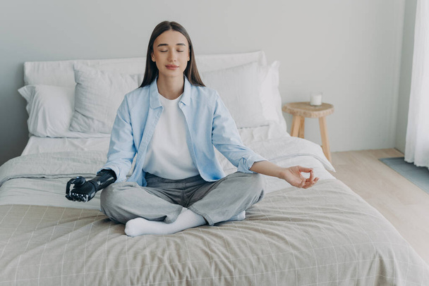 Young disabled woman is sitting in lotus pose on bed with her eyes closed. Attractive european girl has cyber prosthesis. Modern artificial limb. Handicapped woman lifestyle concept. - Foto, immagini