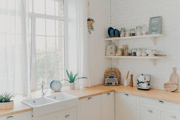 Brightly lit scandinavian kitchen interior. Large window in front of sink. Clean wooden counters along the walls. Accessories and pot plants. Concept of mortgage and moving to modern apartment. - 写真・画像
