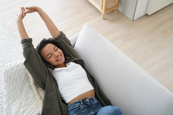 Happy pretty biracial girl resting lying on sofa at home with closed eyes, stretching body. Modern young female waking up after sleep, smiling, enjoying lazy weekend. Domestic life, wellness. - Photo, Image