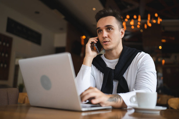 Egy férfi ül az asztalnál a kávézóban vagy az étteremben viseljen alkalmi mobiltelefon, hogy egy hívást beszélő kommunikáció, miközben tart egy csésze kávét valós emberek másolja a helyet - Fotó, kép