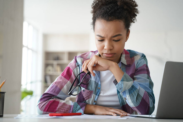 Frustrated afro girl is overworked studying remote at home. Teenage hispanic schoolgirl is sitting at the desk in front of laptop tired from homework. Stressed student has problems with exam. - Φωτογραφία, εικόνα